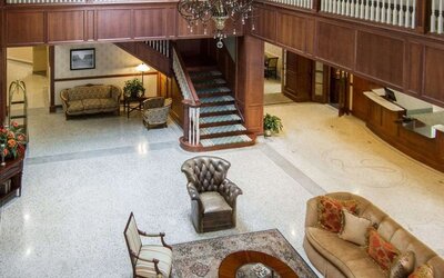Hotel lobby lounge featuring leather armchairs, upholstered sofas, wooden furniture, a decorative chandelier, a central staircase, and a polished terrazzo floor at Liberty Hotel Cleburne, an Ascend Hotel Collection Member.