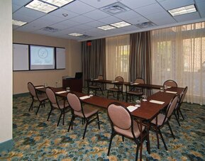 Meeting room arranged with conference tables and chairs in a U-shaped layout, a projection screen, presentation equipment, large curtained windows, and patterned carpeting at Liberty Hotel Cleburne, an Ascend Hotel Collection Member.