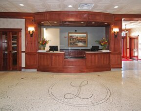 Hotel reception desk featuring rich wood paneling, two computer workstations, floral arrangements, decorative lighting, and a polished floor with an ornamental hotel emblem at Liberty Hotel Cleburne, an Ascend Hotel Collection Member.