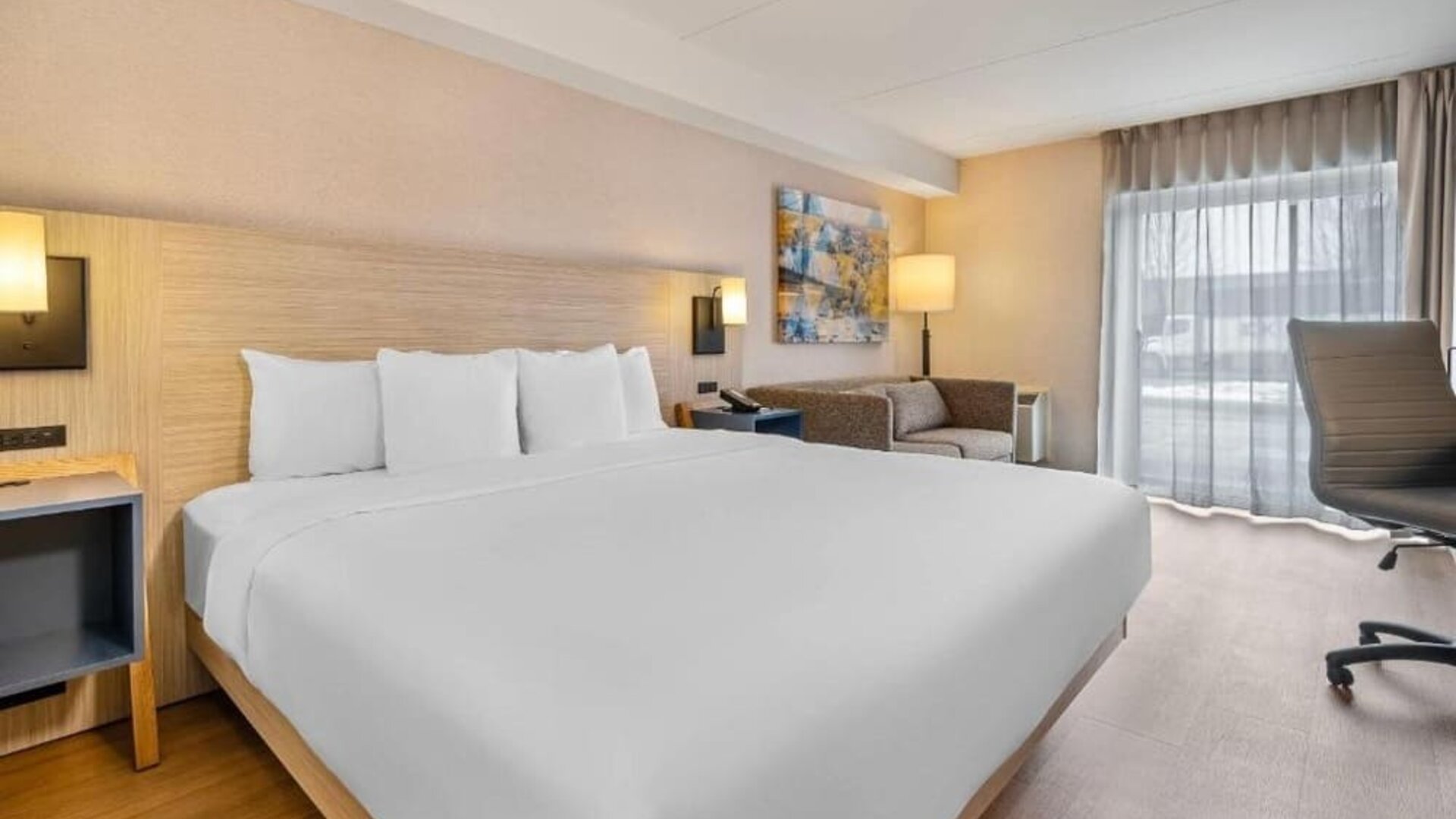 King guest room featuring a large bed with crisp white linens, a light wood headboard, modern wall lighting, blue bedside shelving, a sofa, and contemporary artwork at Comfort Inn Alma.