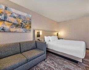 Spacious king guest room with a large bed, crisp white bedding, a gray sofa, abstract wall art, and patterned carpeting at Comfort Inn Alma.