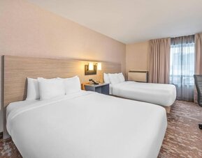 Guest room with two beds featuring white bedding, light wood furnishings, modern wall sconces, a desk and office chair, and neutral decor at Comfort Inn Alma.