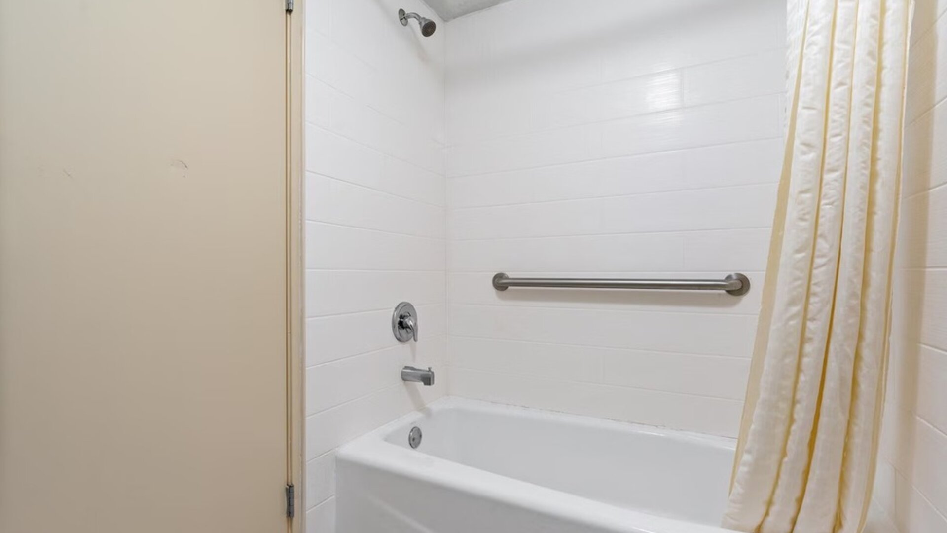 Bathroom featuring a white bathtub and shower curtain, white tiled walls, chrome shower fixtures, and a horizontal stainless-steel grab bar at Chicago Club Inn & Suites.