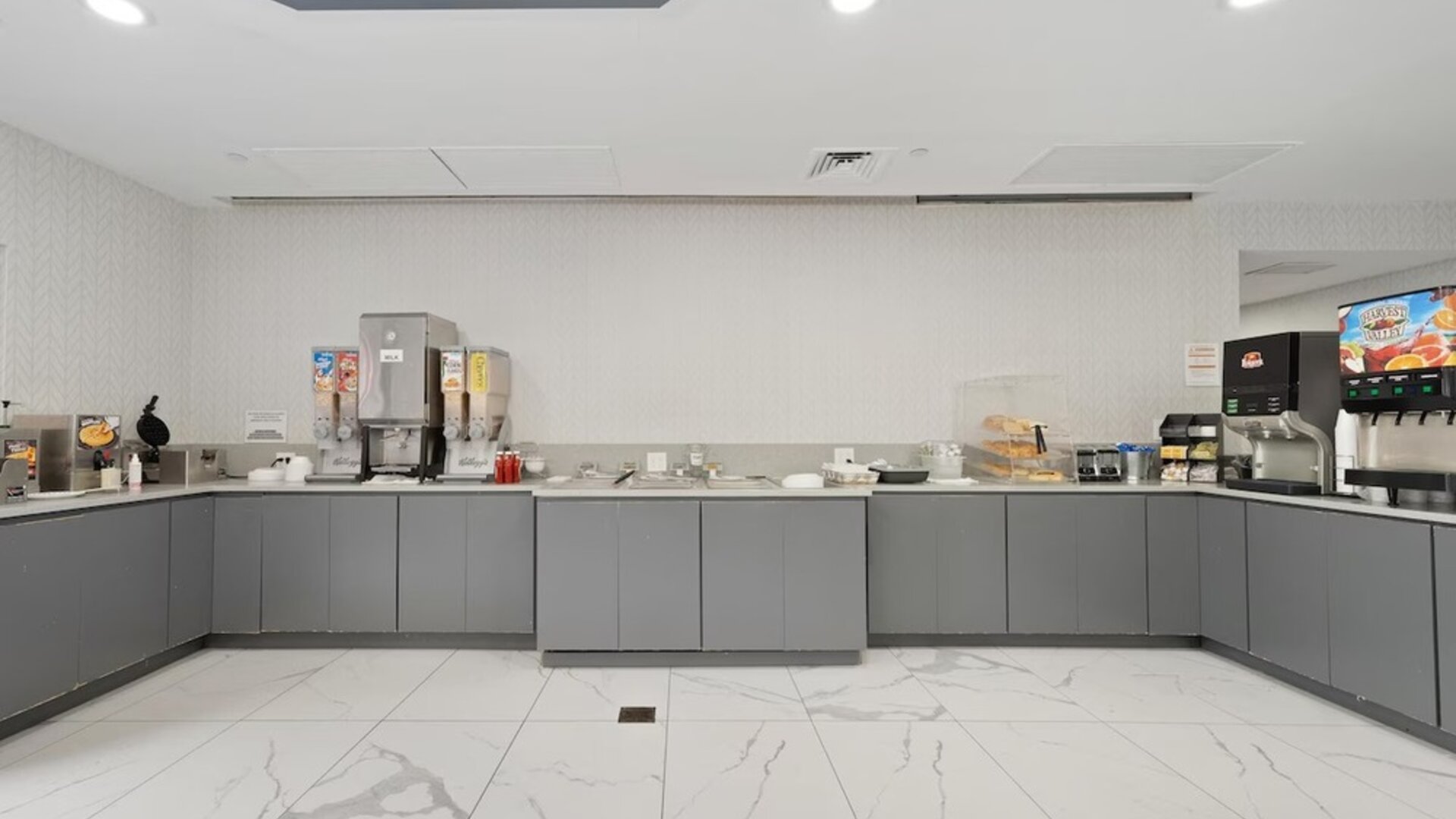 Breakfast buffet area with gray cabinetry, cereal dispensers, beverage machines, serving counters, and a white marble-patterned tile floor at Chicago Club Inn & Suites.