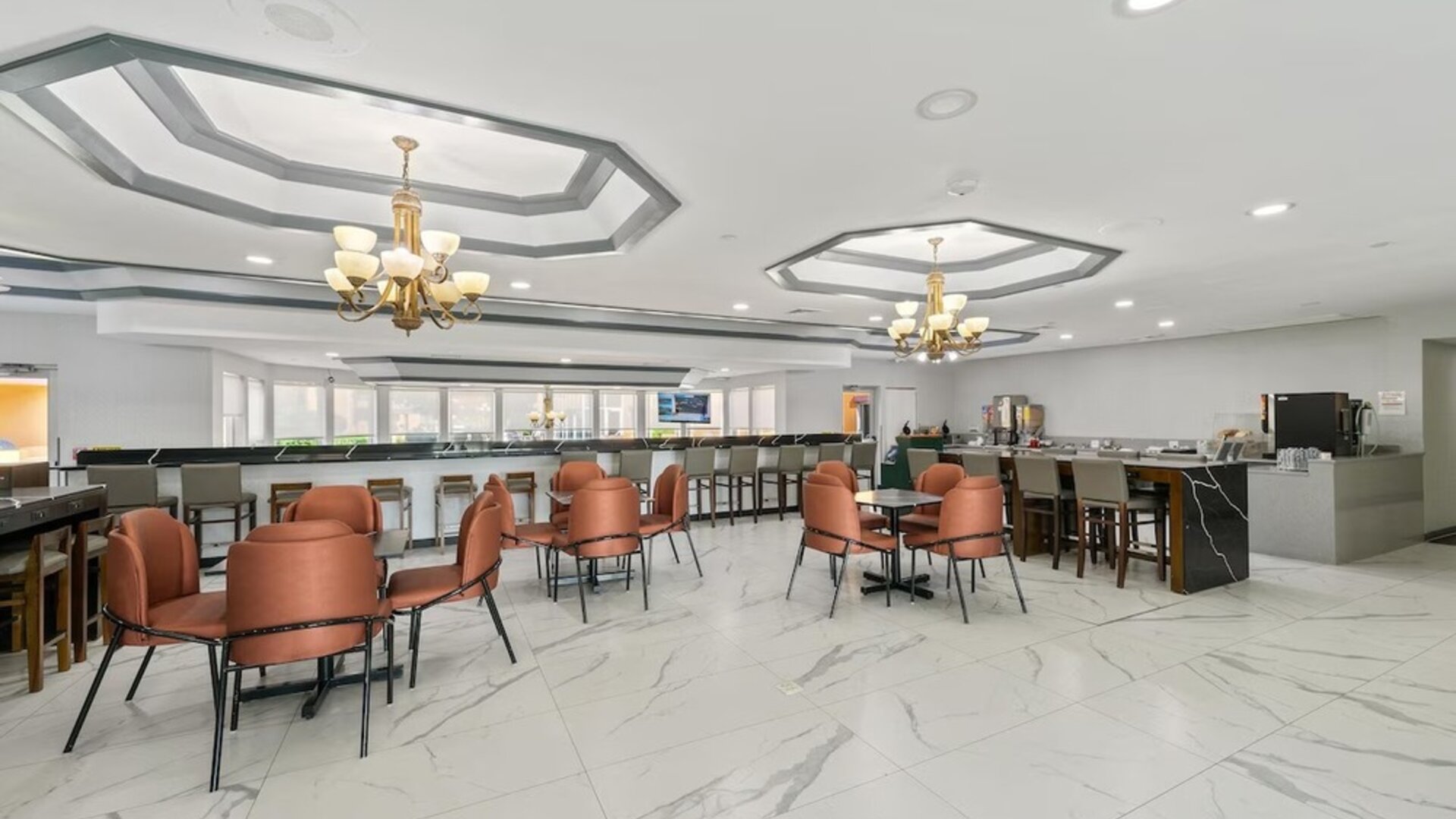 Hotel dining and breakfast area with marble-patterned flooring, orange upholstered chairs, tables, a breakfast counter, and beverage stations at Chicago Club Inn & Suites.