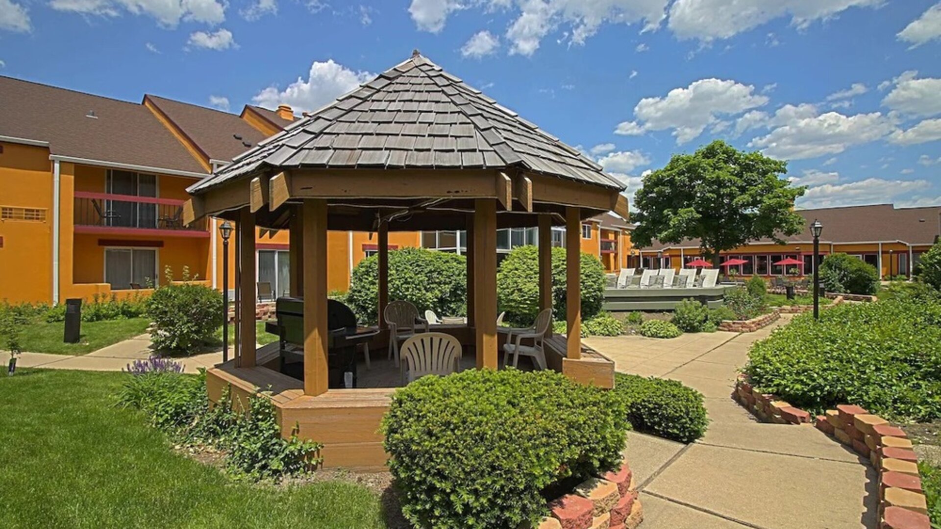 Exterior courtyard featuring a wooden gazebo, landscaped gardens, paved walkways, outdoor seating, and the hotel's orange-colored buildings at Chicago Club Inn & Suites.