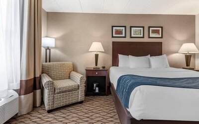 A king bed with white linens and a blue runner beside a patterned armchair, bedside tables and lamps, with a window air-conditioning unit at Comfort Inn & Suites Glen Mills – Concordville.