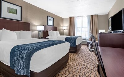 A guest room features two beds with white linens and blue patterned runners, alongside a television, desk and office chair at Comfort Inn & Suites Glen Mills – Concordville.