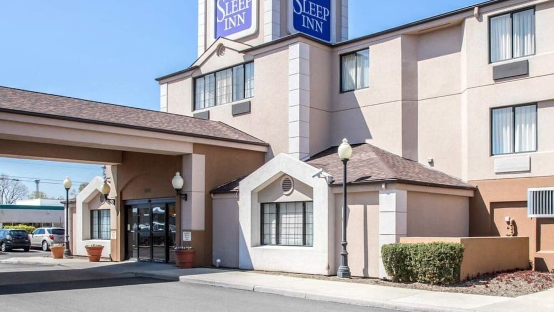 Daytime exterior view of the Sleep Inn hotel featuring a multi-story beige building, prominent blue Sleep Inn signage, covered entrance with glass doors, decorative exterior lighting, landscaped shrubs, and a paved driveway.