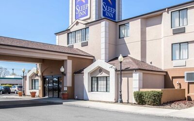 Daytime exterior view of the Sleep Inn hotel featuring a multi-story beige building, prominent blue Sleep Inn signage, covered entrance with glass doors, decorative exterior lighting, landscaped shrubs, and a paved driveway.