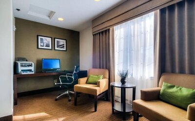 Cozy hotel business center featuring a desktop computer, printer, dark wooden workstation, ergonomic office chair, two beige upholstered armchairs with green accent pillows, a round side table, and a large window providing natural light.
