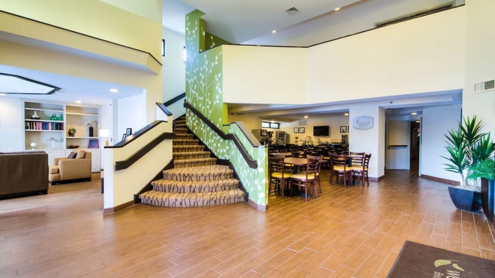 Spacious Sleep Inn lobby featuring a central carpeted staircase with green floral-patterned accent walls, wood-look tile flooring, comfortable lounge seating, an adjacent dining area with wooden tables and chairs, and bright overhead lighting.