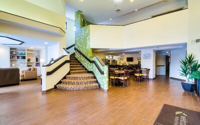 Spacious Sleep Inn lobby featuring a central carpeted staircase with green floral-patterned accent walls, wood-look tile flooring, comfortable lounge seating, an adjacent dining area with wooden tables and chairs, and bright overhead lighting.