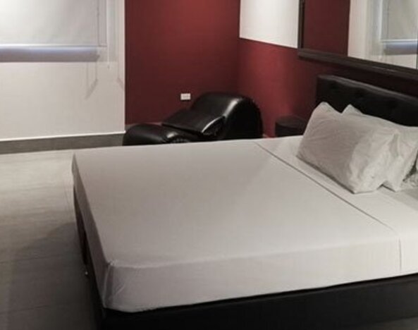 A guest room with a double bed, white linens, a black upholstered headboard, burgundy walls, tiled flooring and a black lounge chair at Residencial & Hotel Palazzo.