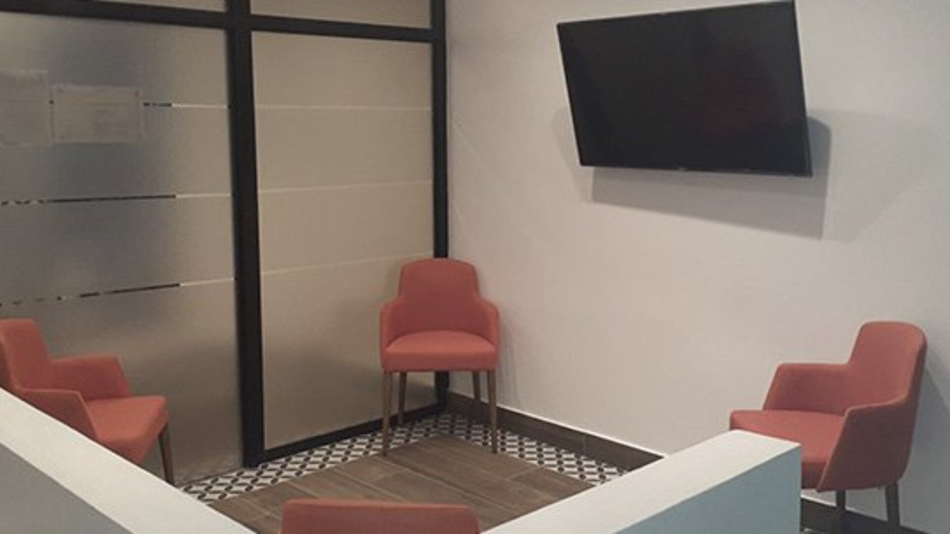 A small lounge area with coral-coloured chairs, a wall-mounted television, tiled flooring and glass partition doors at Residencial & Hotel Palazzo.