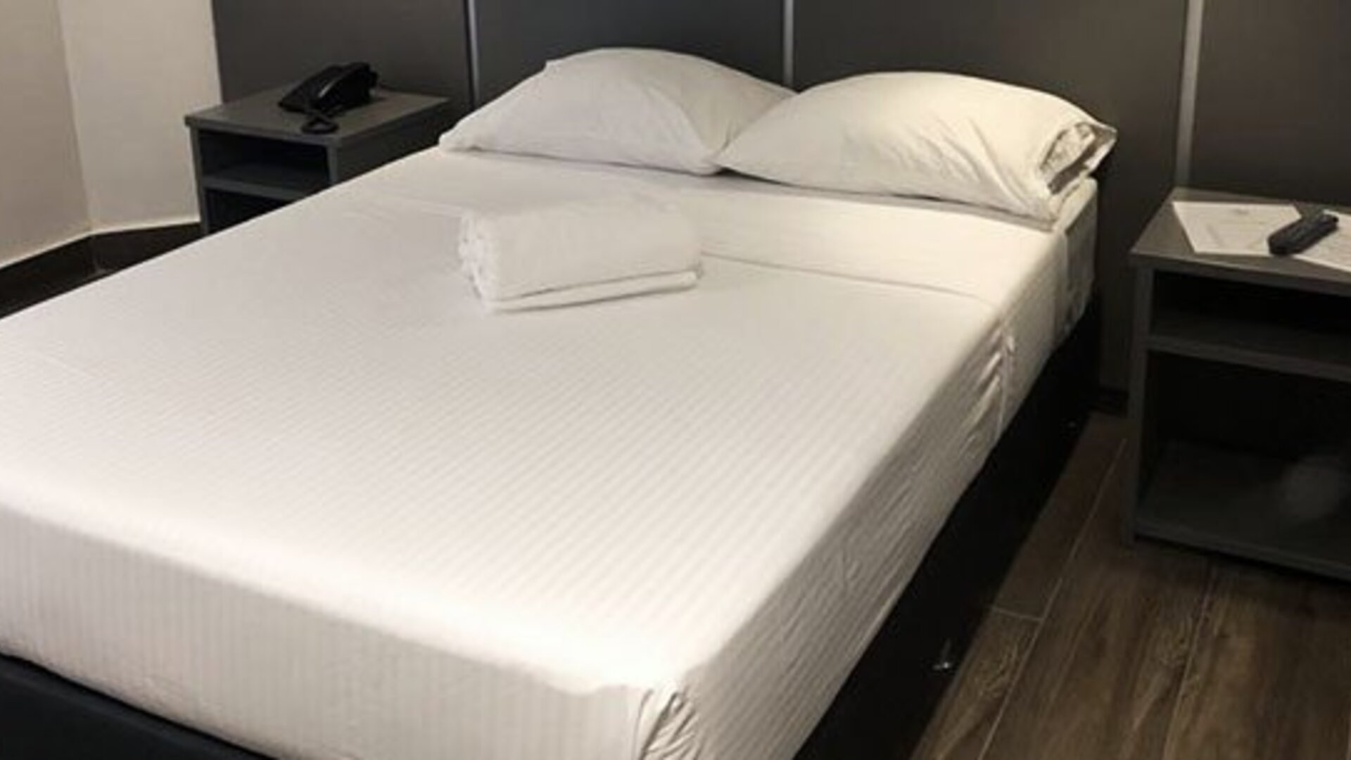 A double bed with white linens and folded towels, dark bedside tables and a black headboard against a dark accent wall at Residencial & Hotel Palazzo.