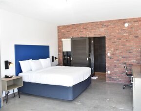 A spacious guest room with a king bed, white linens, a blue upholstered headboard, exposed brick accent wall, desk and office chair at Hotel Bella.