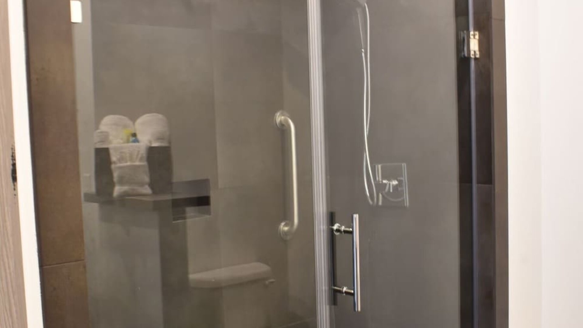 A modern bathroom with a glass-enclosed walk-in shower, dark grey wall tiles, chrome fixtures and a built-in shelf holding folded towels at Hotel Bella.