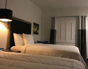 A spacious guest room with two beds, white linens, dark bed frames, a television and a compact kitchenette with white cabinetry at Hotel Bella.