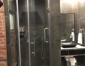 A walk-in shower with glass doors, dark grey tiles, a rainfall showerhead, handheld shower fixture and a wall-mounted shelf at Hotel Bella.