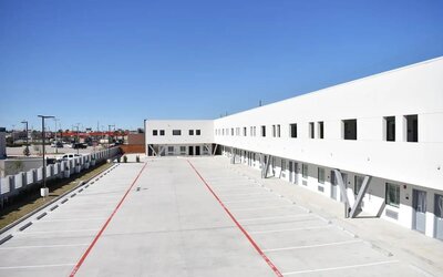 A wide exterior view of the modern white hotel building, showing the open-air corridors, numbered rooms and large concrete parking area at Hotel Bella.