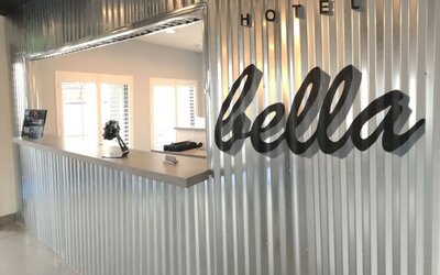 A hotel reception desk set against a corrugated metal wall, featuring a large script-style hotel sign and a bright, minimalist reception area at Hotel Bella.