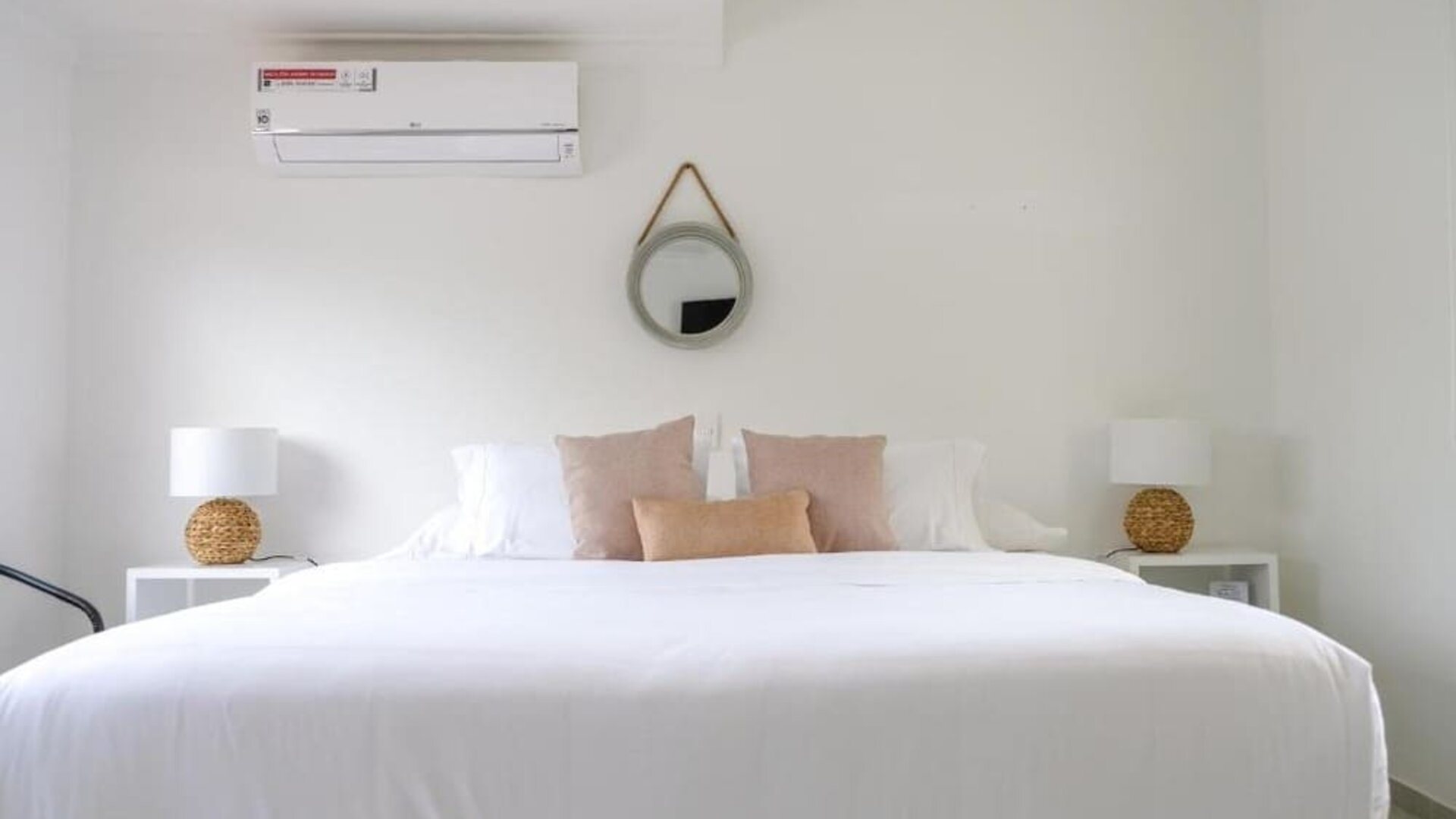 A spacious king bedroom with crisp white bedding, blush and beige accent cushions, matching bedside tables with woven lamps, a round wall mirror and a wall-mounted air conditioner at Mucura Hotel And Spa.