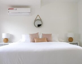 A spacious king bedroom with crisp white bedding, blush and beige accent cushions, matching bedside tables with woven lamps, a round wall mirror and a wall-mounted air conditioner at Mucura Hotel And Spa.