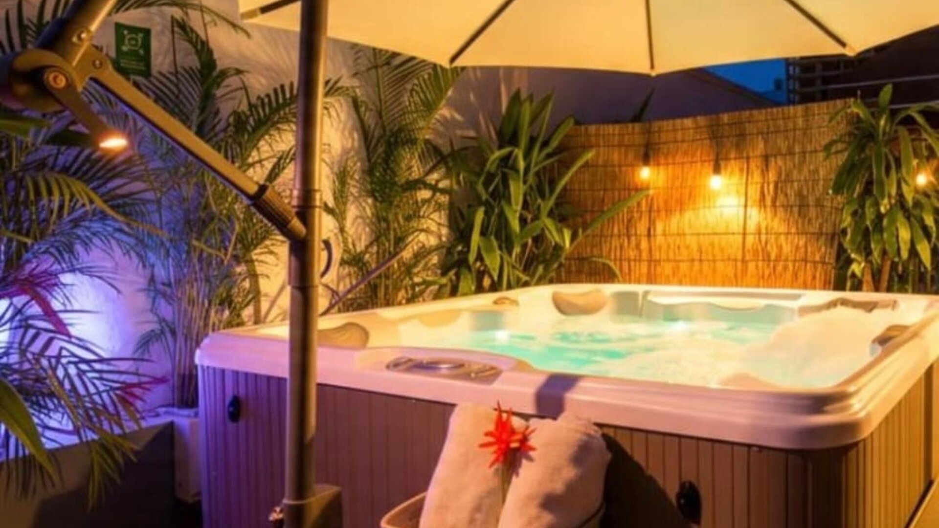An outdoor hot tub surrounded by tropical plants, privacy screening and large cream umbrellas, with ambient lighting creating a relaxing evening atmosphere at Mucura Hotel And Spa.