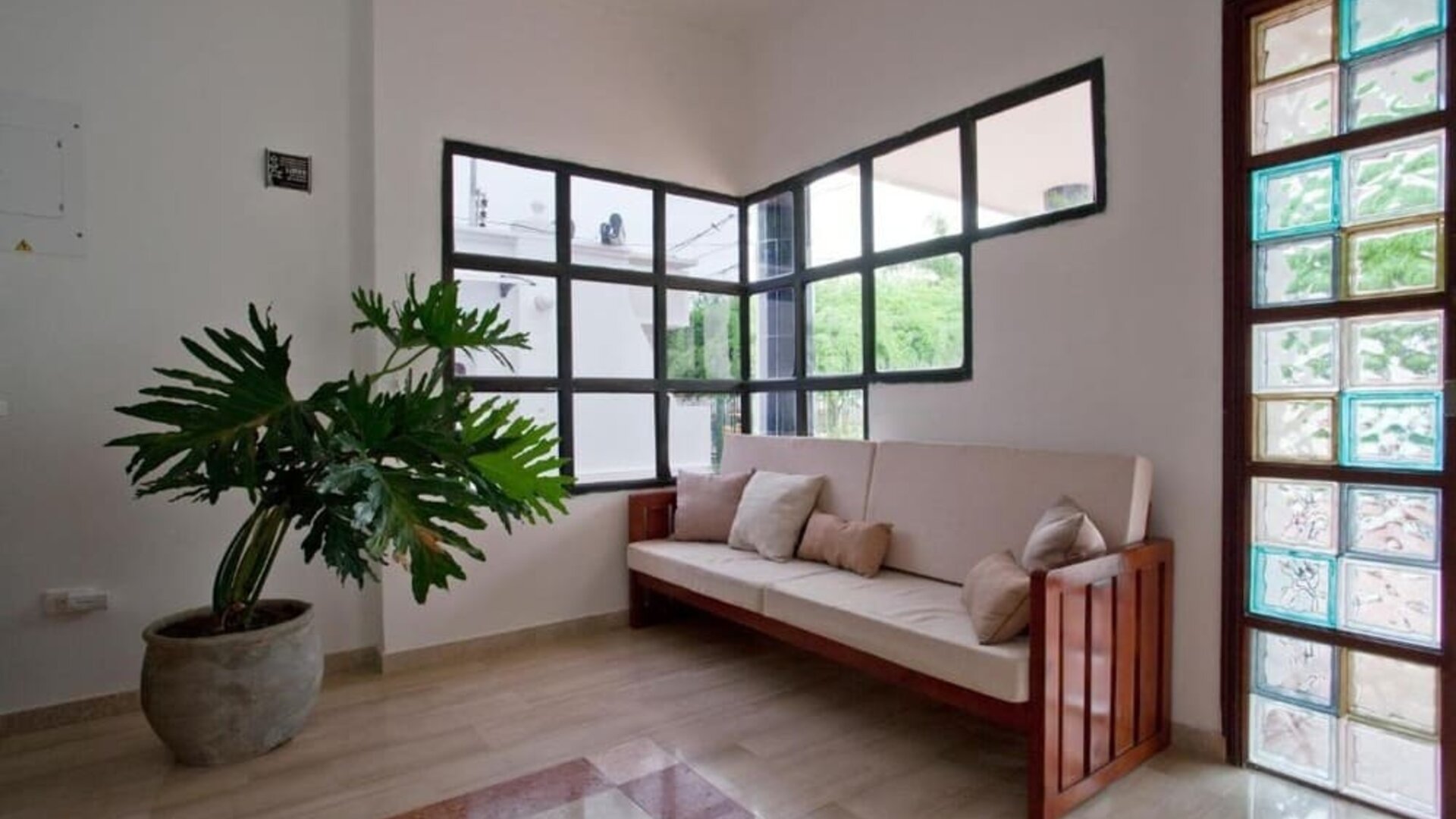 A welcoming lounge area with a wooden-framed sofa upholstered in light fabric, neutral cushions, a large potted plant, tiled flooring and decorative glass-panelled windows at Mucura Hotel And Spa.