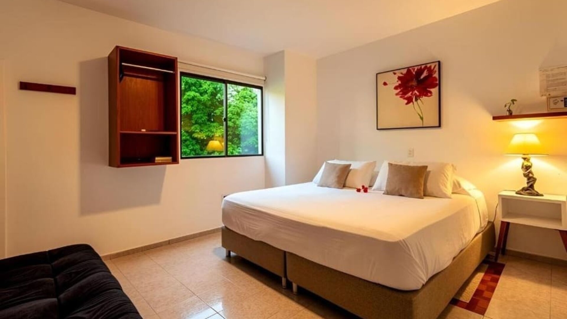 A spacious guest room featuring a king-size bed with white linens and beige cushions, a black sofa, a wall-mounted television, wooden shelving and a large window overlooking greenery at Mucura Hotel And Spa.
