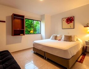 A spacious guest room featuring a king-size bed with white linens and beige cushions, a black sofa, a wall-mounted television, wooden shelving and a large window overlooking greenery at Mucura Hotel And Spa.