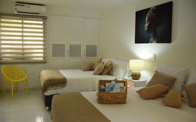 A twin-bedroom setup with two beds dressed in white linens and beige throws, decorative cushions, a yellow accent chair, built-in wardrobes and a wall-mounted air conditioner at Mucura Hotel And Spa.