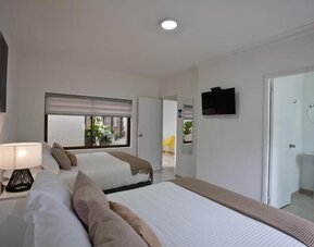 A twin-bed guest room with white bedding, beige accent cushions and throws, a bedside table with a decorative lamp, a wall-mounted television and an en-suite bathroom visible through the open doorway at Mucura Hotel And Spa.