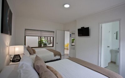 A twin-bed guest room with white bedding, beige accent cushions and throws, a bedside table with a decorative lamp, a wall-mounted television and an en-suite bathroom visible through the open doorway at Mucura Hotel And Spa.