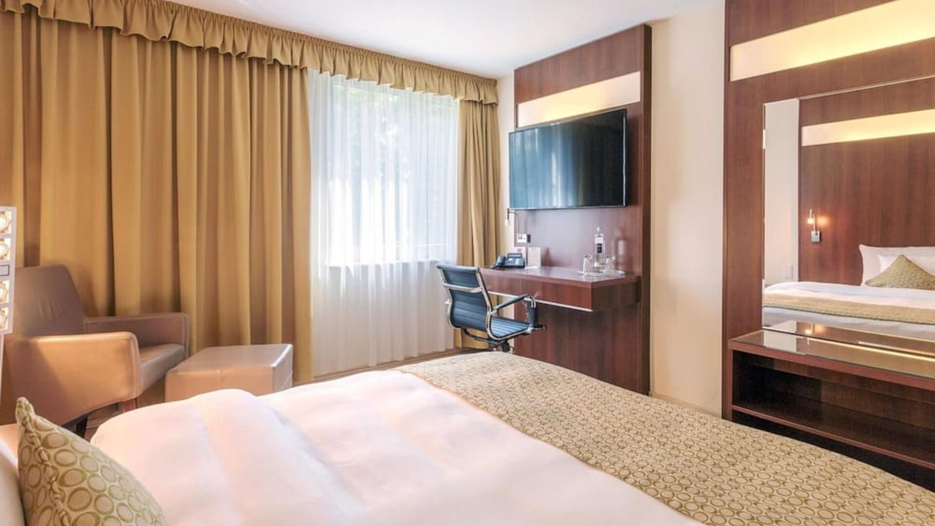 A guest bedroom with a king bed dressed in white linens and patterned bedding, warm wood-panelled furniture, a desk and chair, a large wall-mounted television and a comfortable seating area at Best Western Macrander Hotel Frankfurt/Kaiserlei.