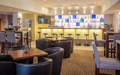 The bar and lounge area at Best Western Macrander Hotel Frankfurt/Kaiserlei, featuring an illuminated backlit bar with a selection of drinks, bar stools, comfortable armchairs, wooden tables and patterned carpeting.