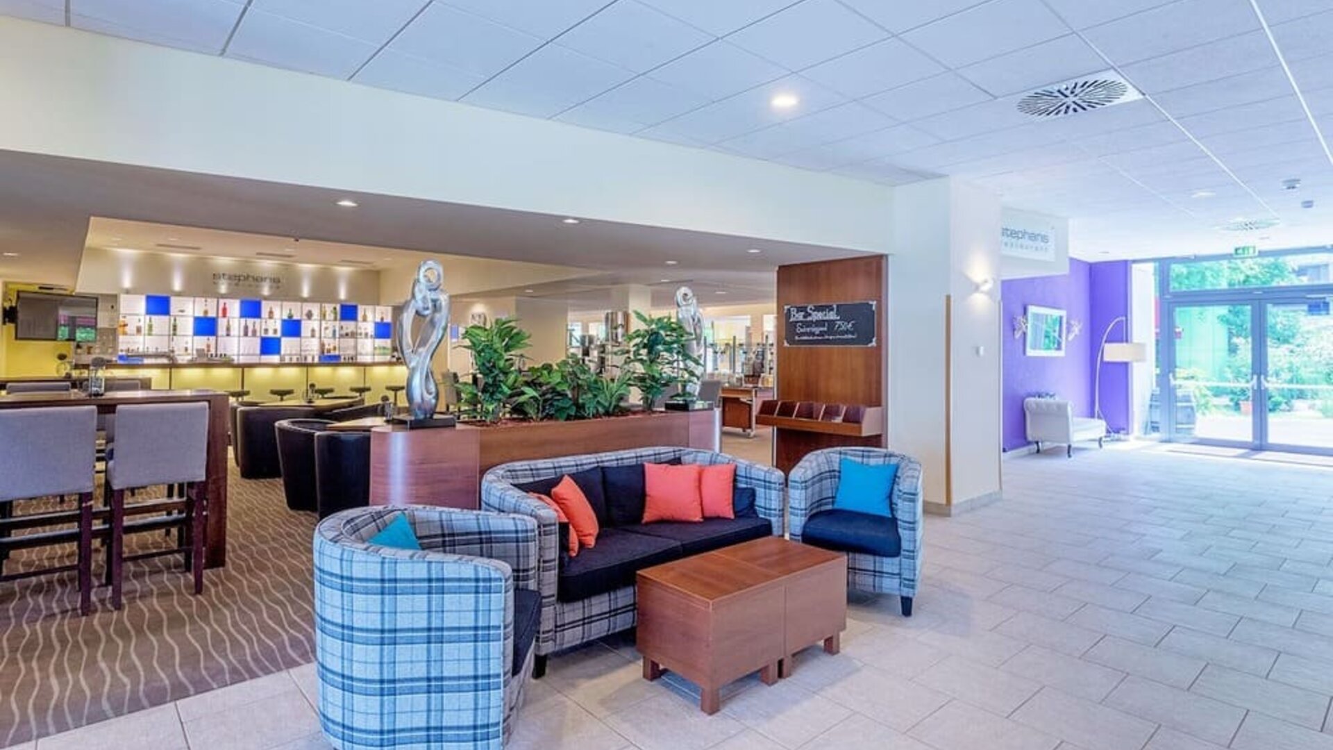 The hotel lobby lounge, featuring a comfortable seating arrangement with plaid armchairs, a sofa with colourful cushions, wooden coffee tables, decorative sculptures and an adjacent bar area.