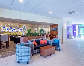 The hotel lobby lounge, featuring a comfortable seating arrangement with plaid armchairs, a sofa with colourful cushions, wooden coffee tables, decorative sculptures and an adjacent bar area.