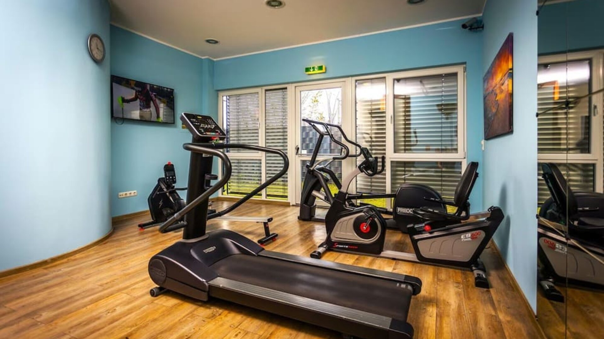 The hotel's fitness room, equipped with treadmills, stationary exercise bikes and other cardio equipment, with mirrored walls, blue accents and wood-effect flooring at Best Western Macrander Hotel Frankfurt/Kaiserlei.