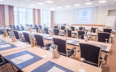 A conference room arranged for meetings and presentations, with rows of tables and chairs, writing materials, water bottles, presentation equipment and a projection screen displaying the Best Western logo.