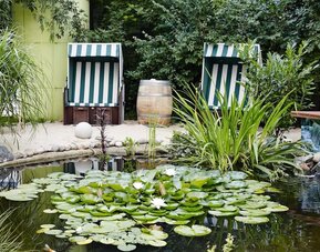 A peaceful landscaped garden with a small pond covered in lily pads and water lilies, surrounded by lush greenery and two striped outdoor deckchairs.