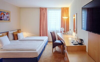 Guest room with two single beds dressed in white linens and patterned bedding, warm-toned curtains, a work desk with a chair, a wall-mounted television and a hospitality tray with a kettle and cups at Best Western Macrander Hotel Frankfurt/Kaiserlei.