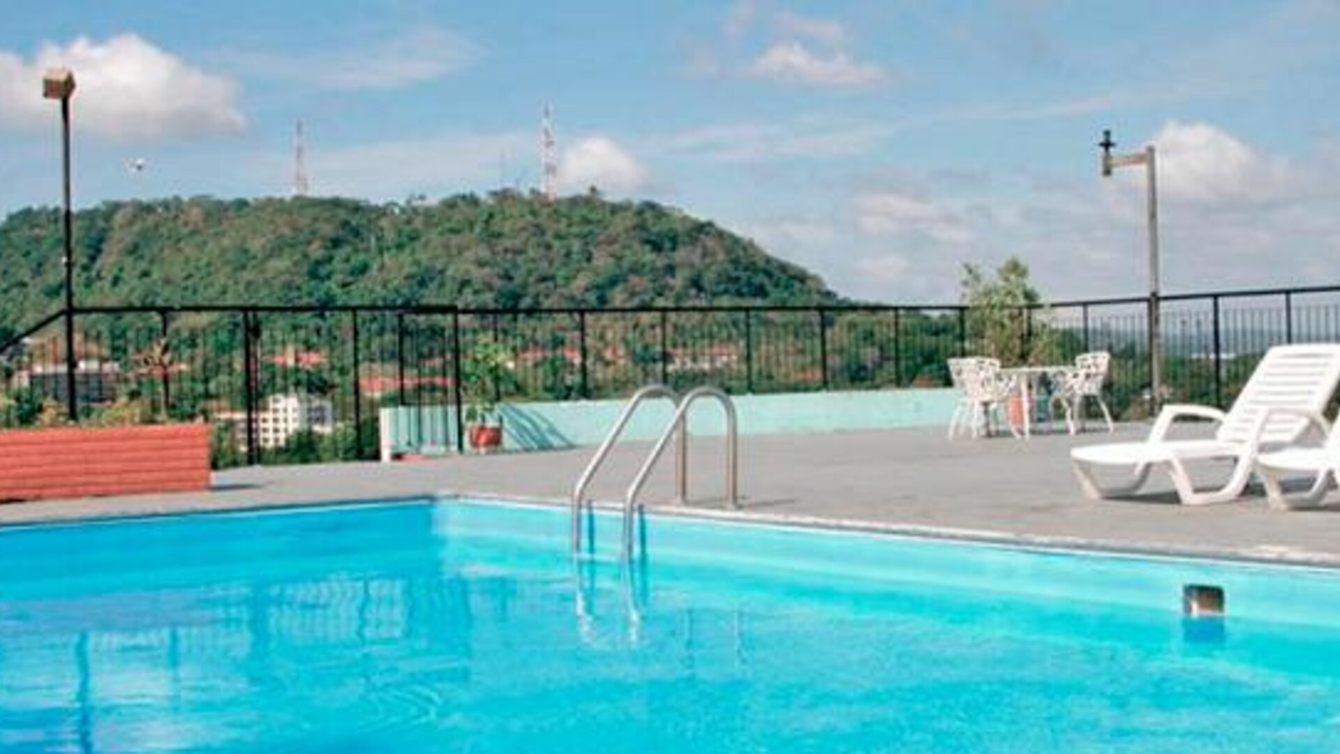 Rooftop swimming pool with white loungers, black railings and a green hillside in the background at Caribe Panamá Hotel.