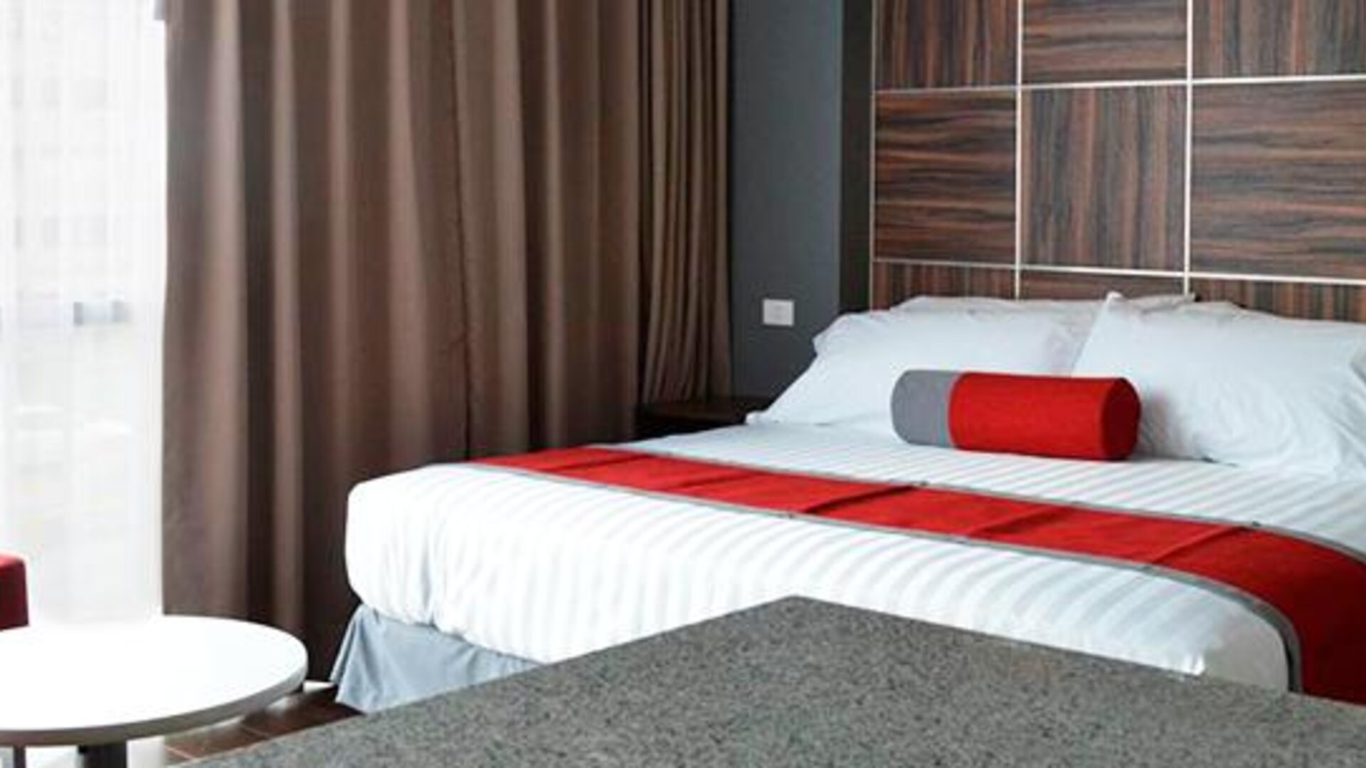 Guest room with a king-size bed, white bedding, red accents, bedside furniture and dark curtains beside a bright window at Hotel Block Suites.