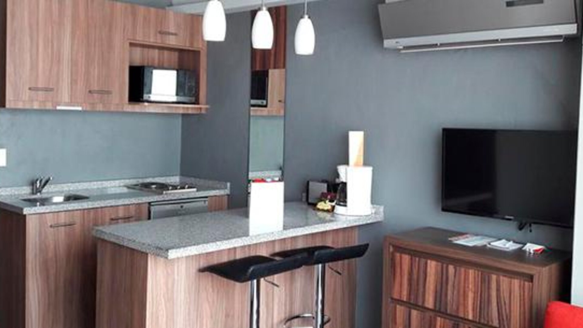 Modern kitchenette and living area with wood cabinetry, microwave, granite breakfast bar, bar stools and wall-mounted television at Hotel Block Suites.