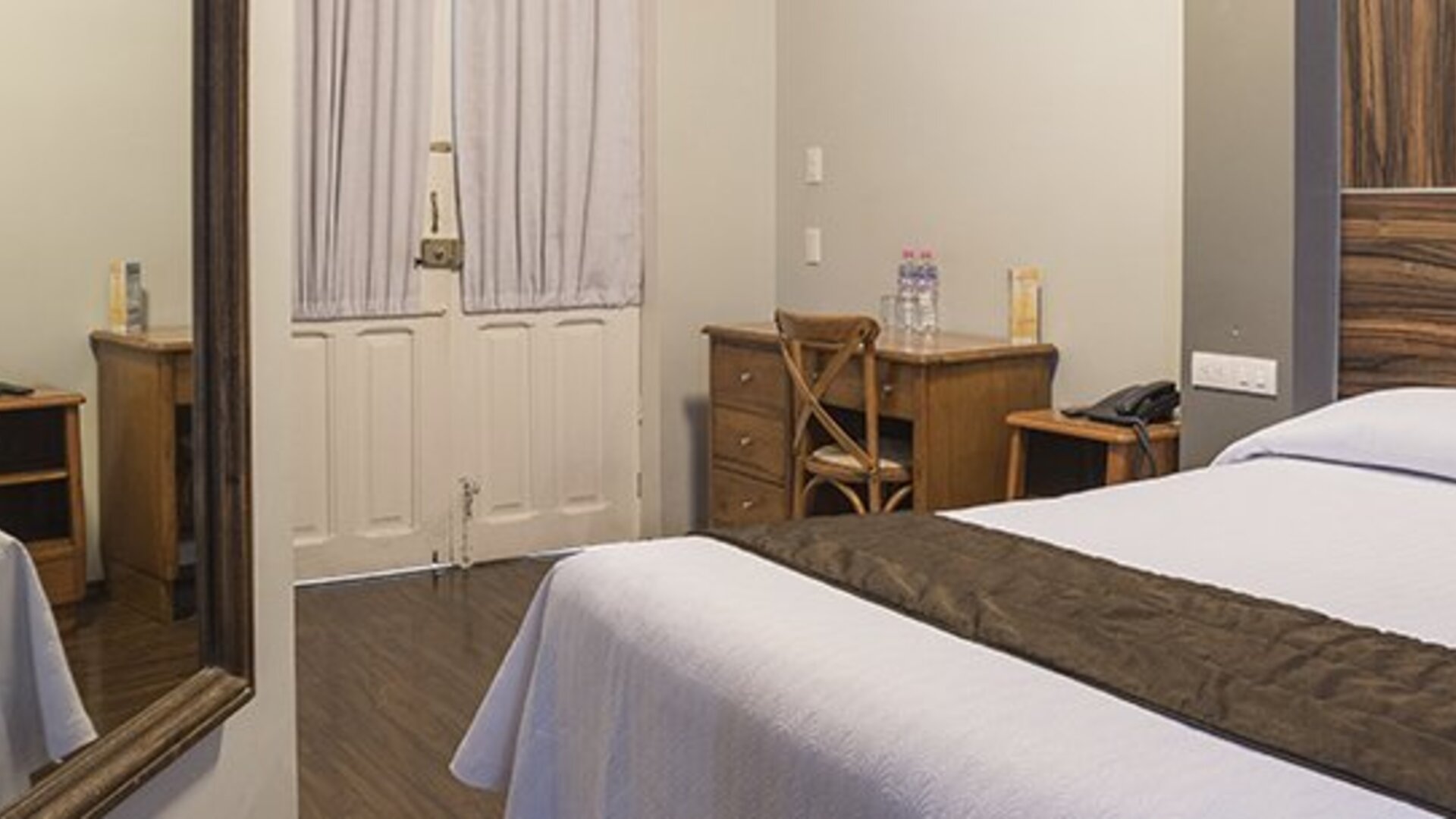 Guest room with a bed beside a wooden desk and chair, with a large wall mirror at Hotel Principal.