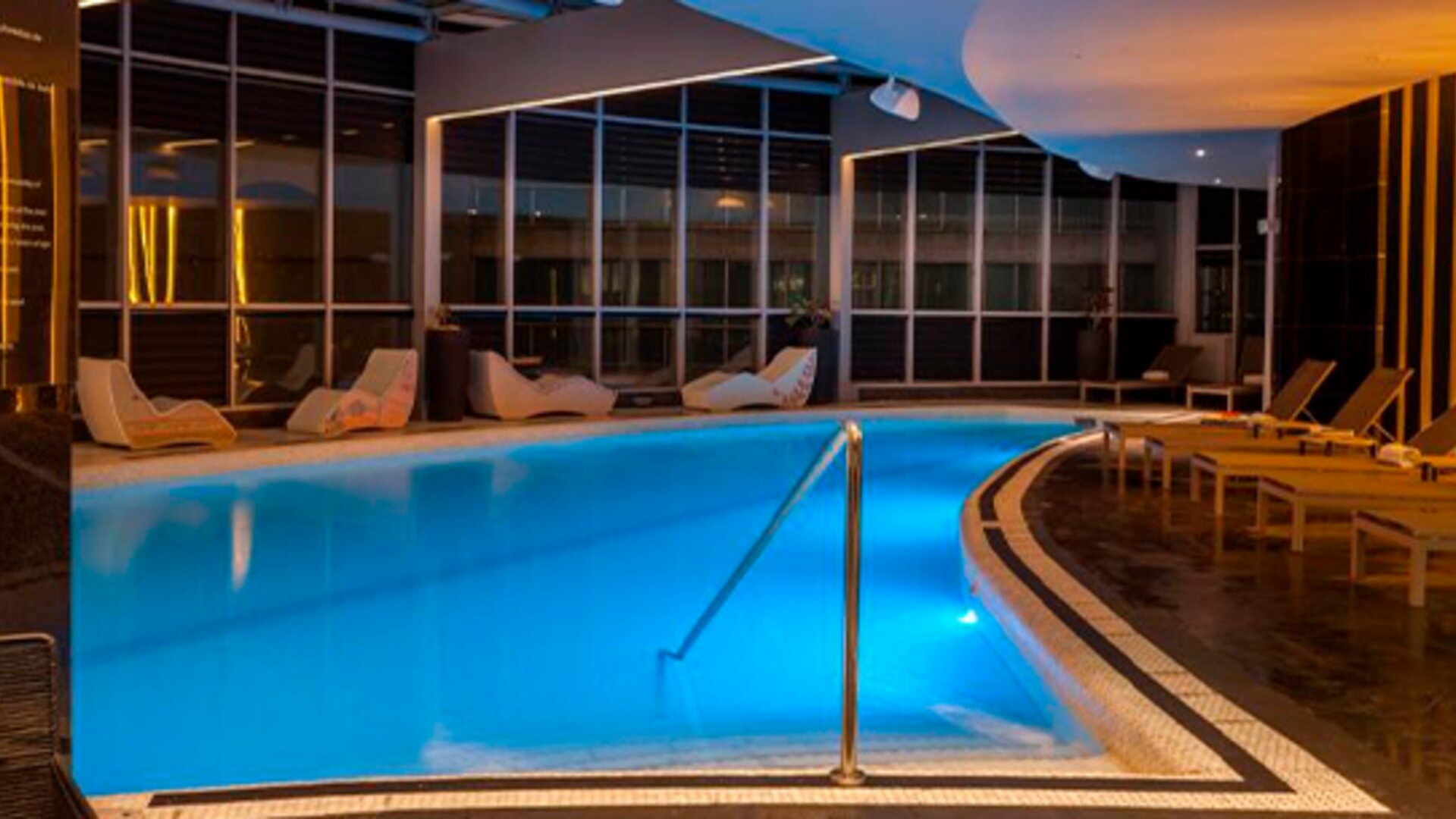 Indoor swimming pool with turquoise water, surrounding loungers and warm ambient lighting at Hotel Las Américas Golden Tower Panamá.