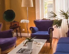 Lounge with purple upholstered sofas and armchairs, a white coffee table, wooden flooring and large windows at Hotel Chemin.