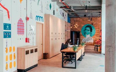 Shared workspace with a long wooden table, chairs, lockers, colourful wall illustrations and exposed concrete columns at UMA Suites Chía.